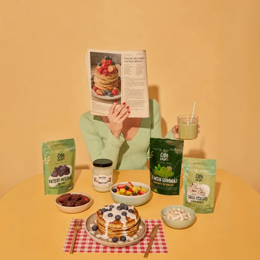 Pancake con mirtilli e crema di cocco su un tavolo giallo, con le buste CiboCrudo di datteri, matcha e cocco